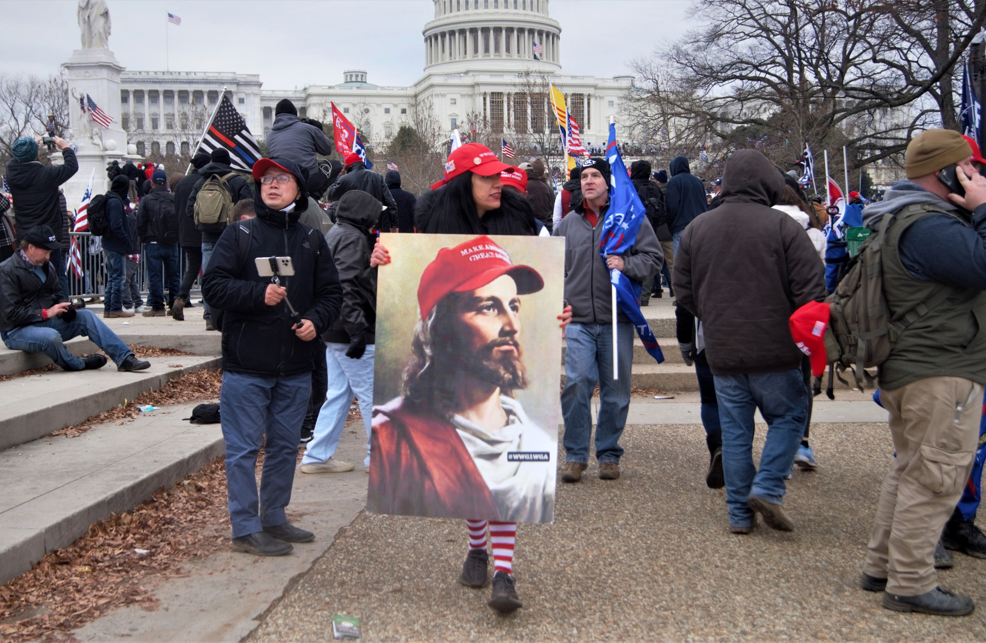 A woman holds a poster with a portrait of Jesus wearing a MAGA hat as other flags and signs wave in the background